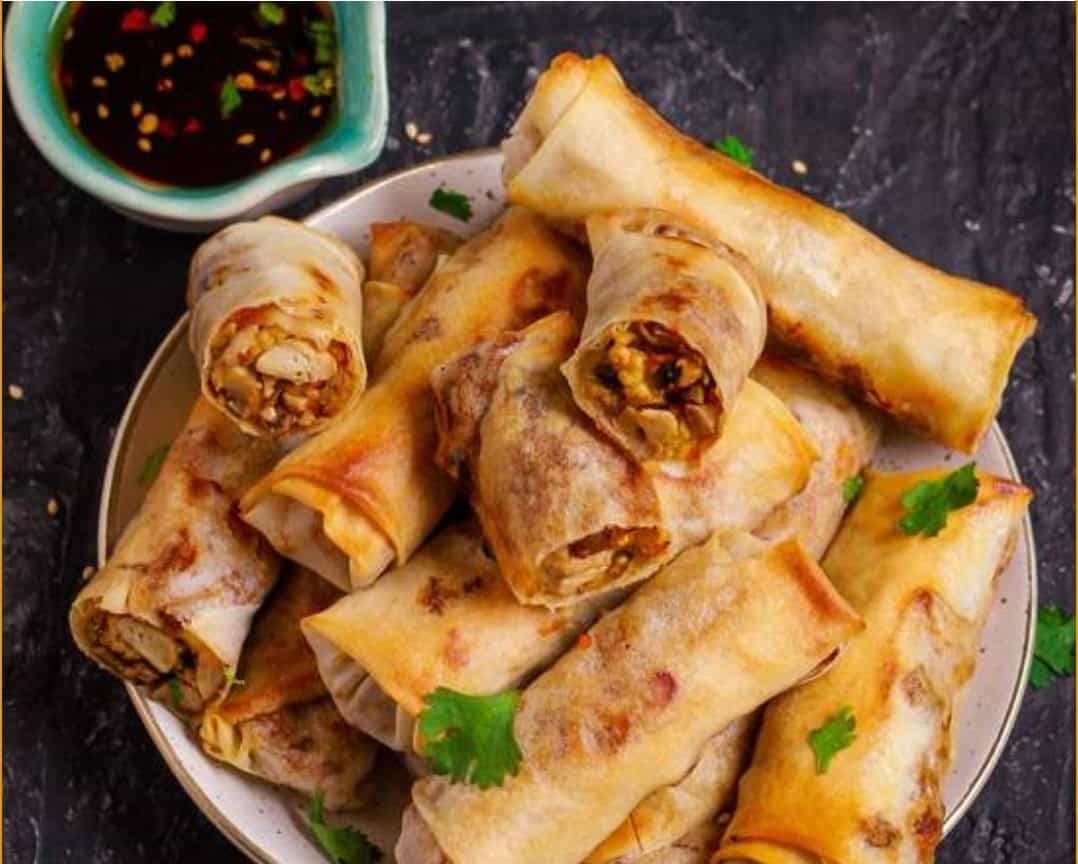 Crispy spring rolls filled with savory vegetables and meat, served with a side of sweet soy dipping sauce, on a dark textured surface garnished with cilantro.