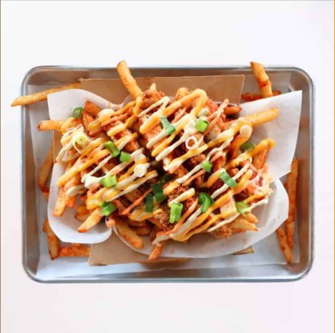 Crispy loaded fries topped with drizzled sauces, green onions, and crispy chicken pieces served in a stainless steel tray, perfect for sharing or enjoying as a flavorful snack.