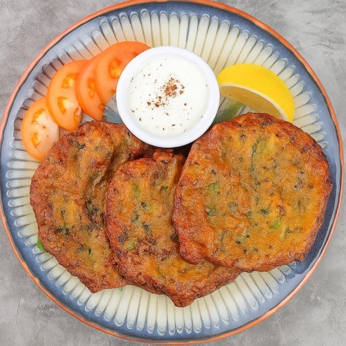 Crispy vegetable cutlets served with fresh tomato slices, lemon wedge, and a side of yogurt-based dip on a decorative ceramic plate. Perfect for healthy snack or appetizer.